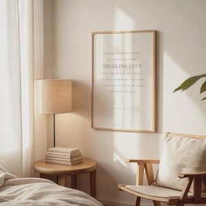 Könnte beinhalten: Ein gerahmter Druck mit hellem Holzrahmen hängt an einer Wand. Der Druck zeigt elegante Typografie mit den Worten "UNFAILING LOVE" prominent dargestellt. Eine Lampe mit beigem Schirm und ein kleiner Beistelltisch aus Holz mit Büchern befinden sich im Vordergrund.