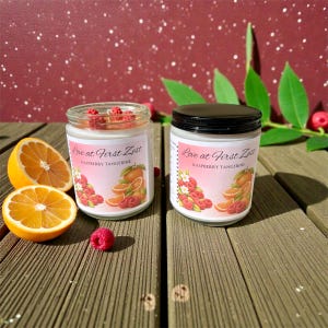 Pode incluir: Dois frascos de vidro de velas perfumadas com tampas pretas, rotulados "Love at First Zest Raspberry Tangerine". Os frascos estão ao lado de fatias de laranja e framboesas. O fundo é vermelho escuro com manchas brancas.