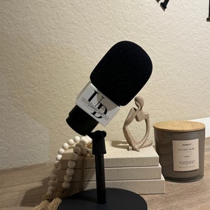May include: A black microphone with a black foam cover and a white label that says "Unfiltered Truth Podcast" is on a black stand. The microphone is on a wooden table with a white beaded garland, a small beige sculpture, and a brown candle with a wooden lid.