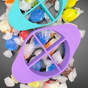 May include: Two plastic candy cutters, one teal and one purple, with four sections each. The cutters are on a black surface with a pile of wrapped candies in various colors.