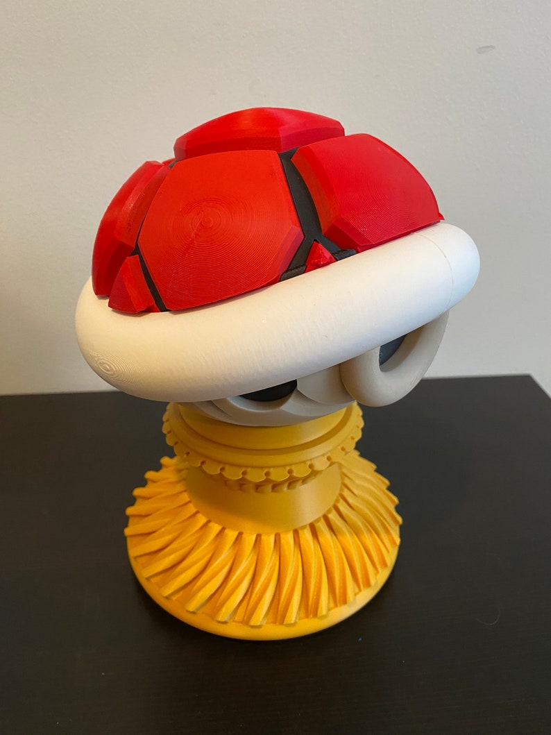 May include: A 3D printed figurine of a red and white Koopa Troopa from the Super Mario Bros. video game series. The figurine is standing on a yellow base.