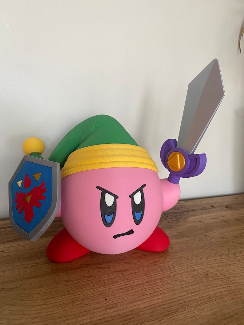 May include: A pink and red 3D printed figurine of a character from the video game Kirby, holding a sword and shield. The character is wearing a green hat with a yellow band.