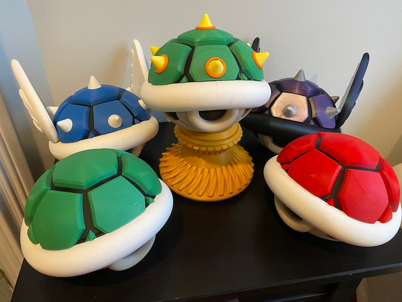 May include: Five different colored turtle shell figurines from the Super Mario Bros. video game franchise. The shells are green, blue, red, black, and yellow. The yellow shell has a single eye and is on a yellow stand.