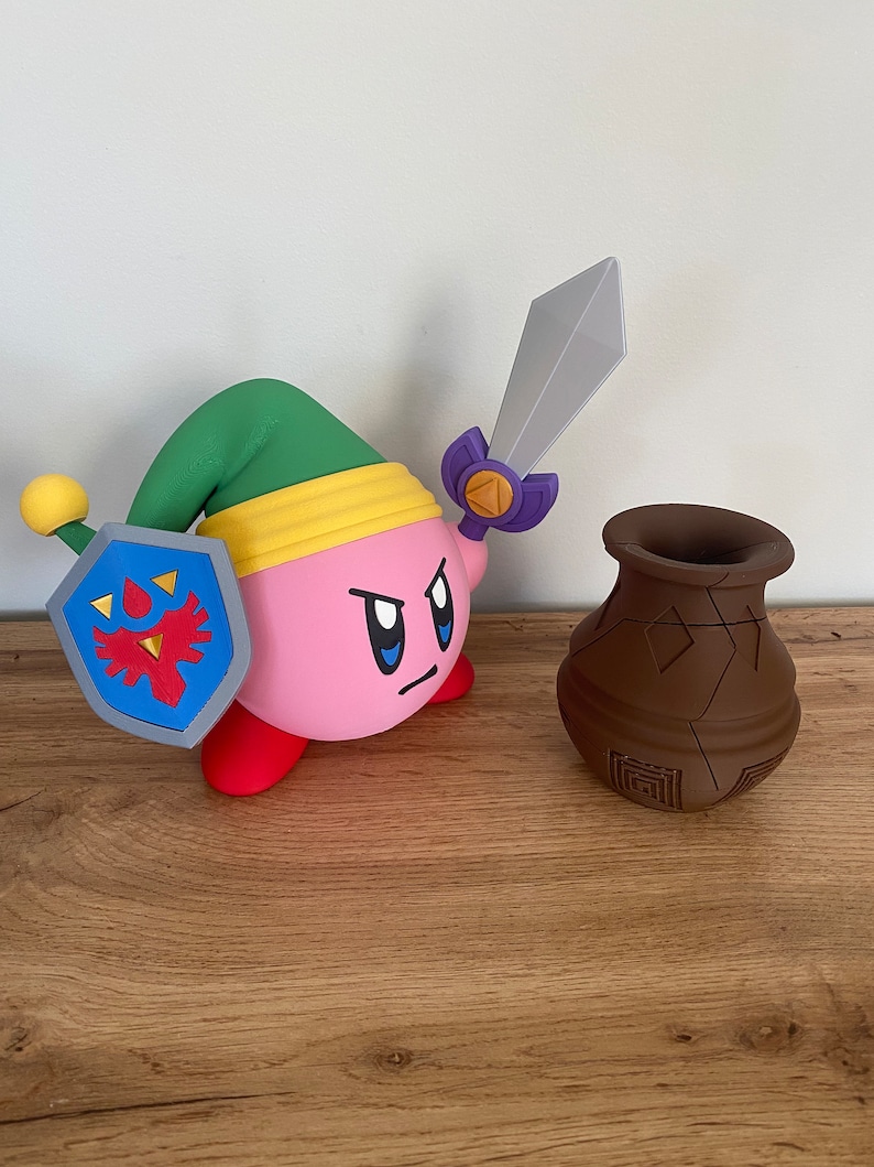 May include: A pink and red character with a green hat and a sword, holding a blue and red shield with a red bird design. The character is standing on a wooden surface next to a brown clay pot.