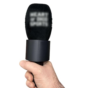 May include: A black microphone with a black foam windscreen is held in a hand. The microphone has a cylindrical base and a handle. The text "HEART OF SPORTS" is visible on the microphone.