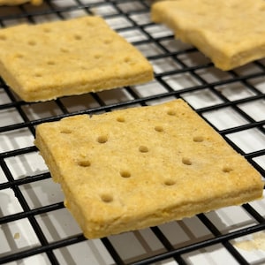 May include: A close-up shot of several golden-brown, square crackers with small holes, arranged on a black wire rack. The crackers have a textured surface and are likely freshly baked. The image suggests a homemade or artisan food product.