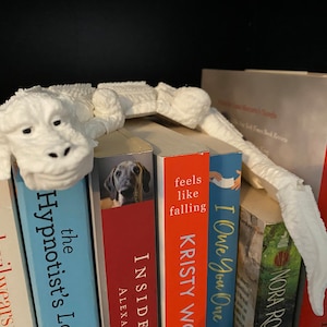 May include: A white, 3D printed bookend shaped like a creature with a long tail, holding a stack of books. The books have titles like "The Hypnotist's Love", "The Devil Wears Prada", "Inside", "Feels Like Falling", "I Owe You One", and "Nora Roberts".