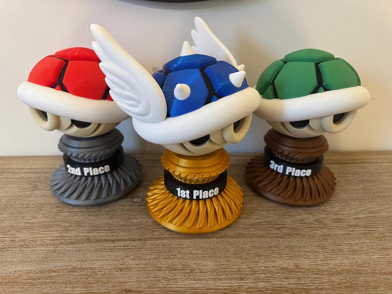 May include: Three 3D printed trophies shaped like a Koopa shell from the Super Mario Bros. video game series. The trophies are painted red, blue, and green and have a white base. The bases are labeled "1st Place", "2nd Place", and "3rd Place".