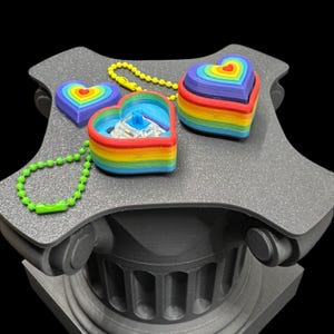 May include: A collection of rainbow-colored heart-shaped trinkets, some with keychains, displayed on a gray, decorative pedestal. The hearts feature layers of vibrant colors, including red, orange, yellow, green, blue, and purple. The background is black.