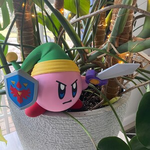 May include: A pink and green 3D printed figurine of a character from the video game Kirby. The character is holding a sword and a shield with a bird design. The figurine is sitting in a pot with a green plant.