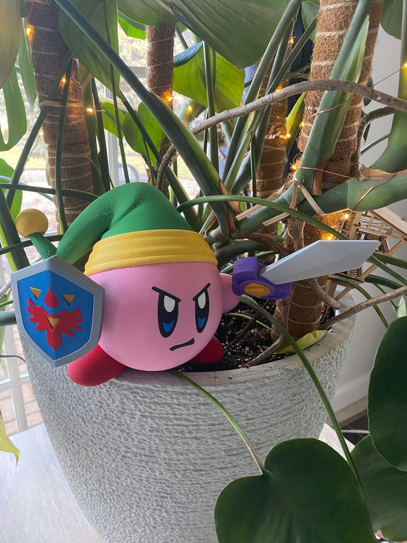 May include: A pink and green 3D printed figurine of a character from the video game Kirby. The character is holding a sword and a shield with a bird design. The figurine is sitting in a pot with a green plant.