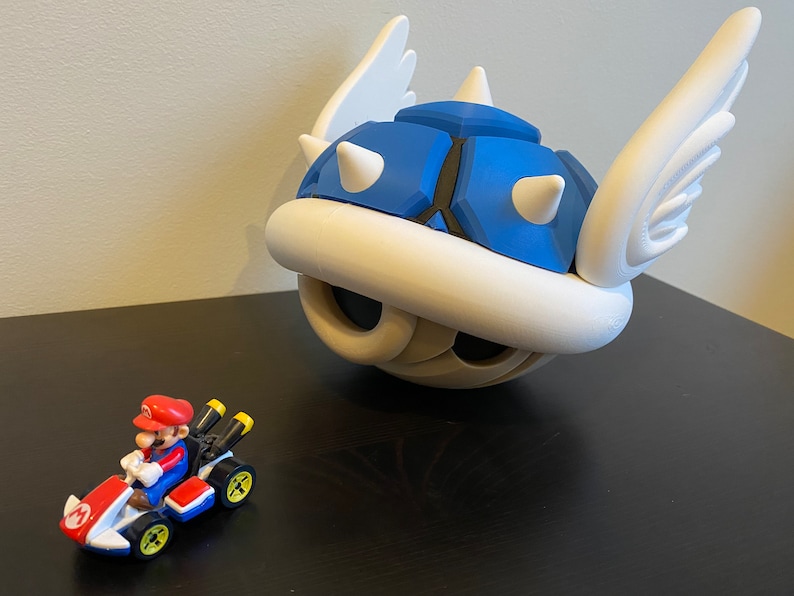 May include: A blue and white winged shell with spikes, a popular character from the Mario Kart video game series, is shown next to a small red and white toy race car with a figure of Mario driving.