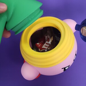 May include: A pink and yellow character with a large yellow rim around its head. The character has a small opening in the top of its head, revealing a variety of chocolate candies inside.