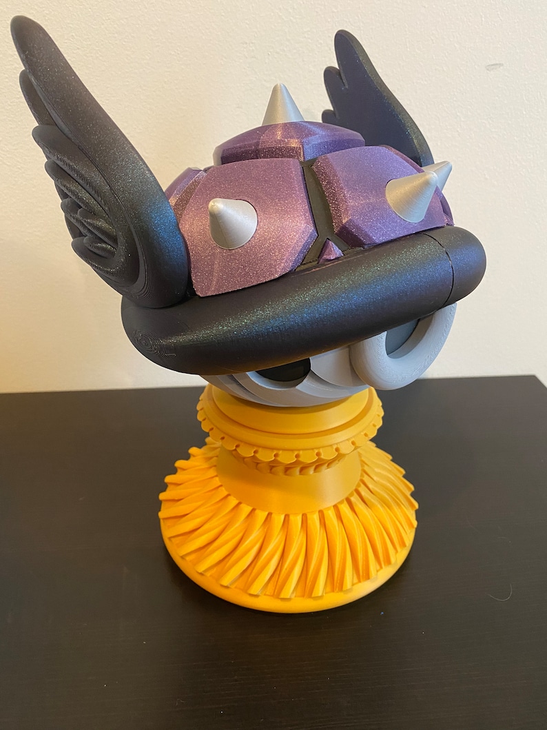 May include: A purple and black 3D printed figurine of a winged Koopa Troopa from the Super Mario Bros. video game series. The figurine is standing on a yellow base.