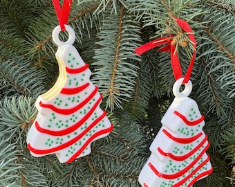Pode incluir: Dois enfeites de árvore de Natal brancos com listras vermelhas e verdes e glitter verde. Os enfeites estão pendurados em uma fita vermelha em um galho de pinheiro verde.