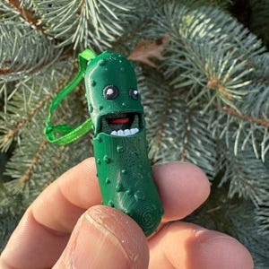 May include: A green pickle-shaped Christmas ornament with a smiling face, white eyes, and red mouth. The ornament has a green ribbon attached to the top.