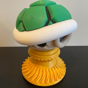 May include: A green and white Super Mario Bros. Koopa shell figurine on a yellow stand. The shell is made of plastic and has a textured surface. The stand is made of plastic and has a decorative design.