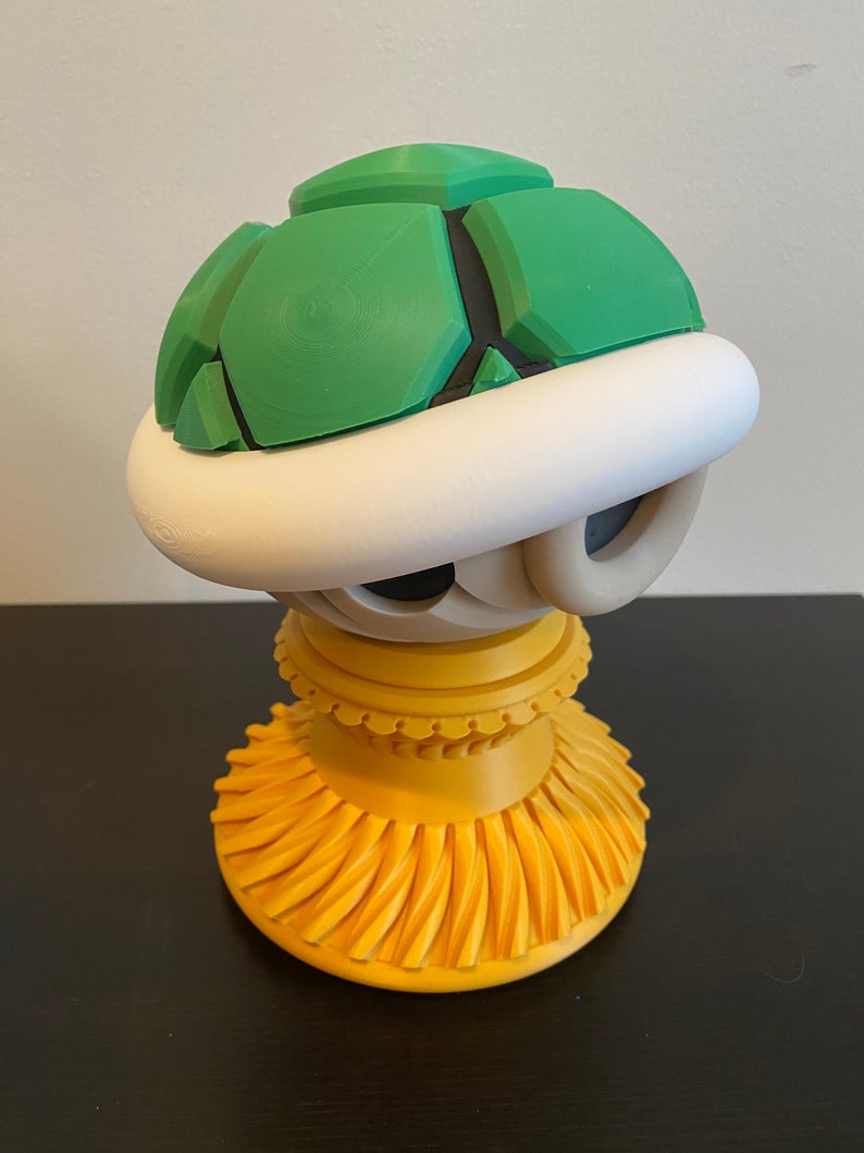 May include: A green and white Super Mario Bros. Koopa shell figurine on a yellow stand. The shell is made of plastic and has a textured surface. The stand is made of plastic and has a decorative design.