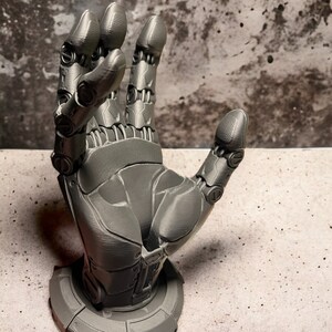 Könnte beinhalten: Eine 3D-gedruckte Roboterhand-Skulptur mit grauer Oberfläche. Die Hand ist in einer Welle oder "Hang Loose"-Geste aufgestellt. Die Hand ist auf einem runden Sockel montiert.