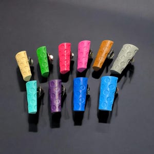 Könnte beinhalten: Sortiment aus zehn bunten, strukturierten, zylindrischen Bleistifthaltern. Jeder Halter hat eine andere Farbe und ein strukturiertes Muster aus kleinen Kreisen. Die Halter sind in zwei Reihen auf einer dunklen Oberfläche angeordnet.