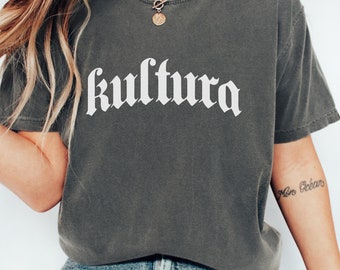 May include: A dark gray t-shirt with the word "kultura" printed in white on the front. The shirt is worn by a person with long brown hair and is styled with blue jeans and white sneakers.