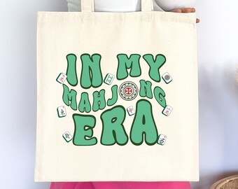 May include: Cream-colored tote bag with the text "IN MY MAH JONG ERA" in green, surrounded by Mahjong tiles. The bag has a rectangular shape and long handles. The design is centered on the bag.