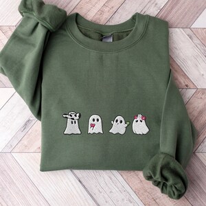May include: Military green sweatshirt with embroidered ghost designs. The ghosts have various accessories, including a hat, a tongue, and a flower crown. The sweatshirt is made of a soft material and is perfect for Halloween or everyday wear.