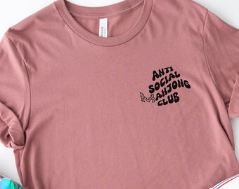 May include: A mauve t-shirt with the text "Anti Social Mahjong Club" in a black, wavy font.