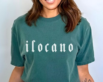 May include: A woman wearing a dark green t-shirt with the word "ilocano" printed in white on the front. She is wearing ripped blue jeans and has a tattoo on her arm.