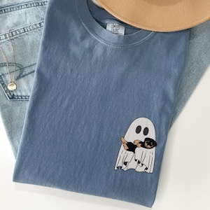 Può includere: Una t-shirt blu jeans con un fantasma bianco e un cane nero e marrone all'interno del fantasma. L'etichetta dice "COMFY COLORS".
