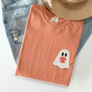 May include: A terracotta orange t-shirt with a white ghost holding a pink donut. The ghost has two black eyes and a small mouth. The t-shirt is folded and laid on top of a pair of blue jeans.
