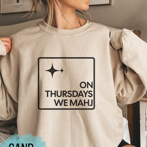 May include: A beige sweatshirt with a black square graphic that says "ON THURSDAYS WE MAHJI" and a star design. The sweatshirt is on a person wearing black shorts.