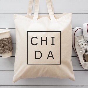 May include: A white canvas tote bag with black handles and the text "CHI DA" printed in a black square on the front. The bag is sitting on a white wooden surface with a cup of coffee and two white trainers.