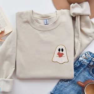 Peut inclure: Un sweat-shirt couleur sable avec un fantôme tenant un cœur brodé sur le devant. Le fantôme est souligné de fil doré.