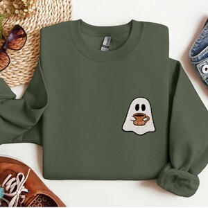 May include: A military green sweatshirt with a white ghost holding a brown coffee cup. The ghost has two black eyes and a small smile.