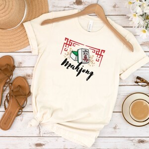 May include: A white t-shirt with a graphic of mahjong tiles and the word "mahjong" in black lettering. The graphic is surrounded by a red and white border.