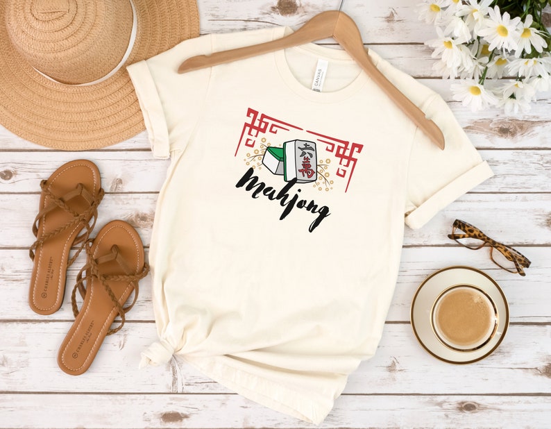 May include: A white t-shirt with a graphic of mahjong tiles and the word "mahjong" in black lettering. The graphic is surrounded by a red and white border.