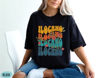 May include: Black t-shirt with a retro-style graphic that says "Ilocano" in colorful letters with flowers and vines.