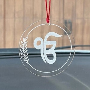 May include: A white acrylic car charm with a red string hanging from it. The charm is round and features a white Khanda symbol with a leafy design around it.