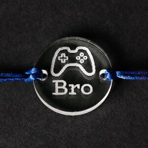 May include: A clear acrylic charm with a white outline of a video game controller and the word "Bro" engraved on it. The charm is attached to a blue cord.