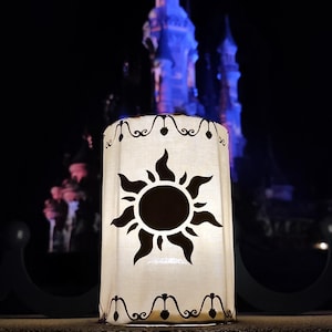 May include: A cylindrical white lantern featuring a black sun graphic with flame-like rays on the front. The top and bottom edges are decorated with a black repeating heart and scroll pattern. The lantern is illuminated from within, glowing brightly.