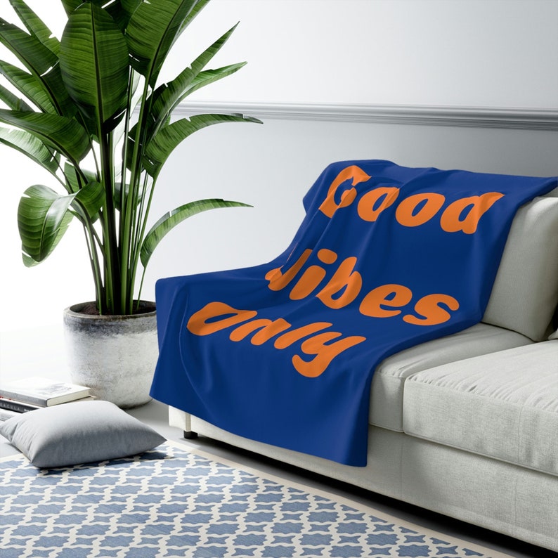 May include: A blue throw blanket with the words "Good Vibes Only" in orange lettering. The blanket is draped over a white couch. A large green plant in a pot is in the background.