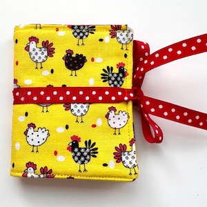 May include: A yellow fabric item with a chicken pattern, tied with a red ribbon with white polka dots. The chicken pattern features white and black chickens with red accents on a yellow background. The ribbon is tied in a bow.