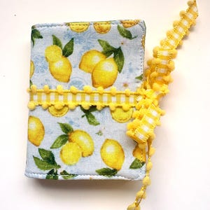 May include: A small, fabric-covered notebook with a lemon pattern. The cover features yellow lemons and green leaves on a light blue background. A yellow pom-pom trim and a yellow and white checkered ribbon are wrapped around the notebook.