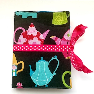 May include: A fabric notebook cover with a black background featuring colorful teapots and teacups. A pink ribbon with white polka dots wraps around the cover and ties on the right side. The teapots are in shades of pink, blue, yellow, and green.