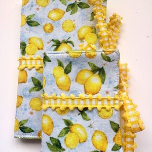 May include: Two fabric items with a lemon pattern on a light blue background. The lemons are yellow with green leaves. Yellow pom-pom trim and a yellow and white checkered ribbon accent the items. The items appear to be a small notebook and a larger item.