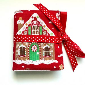 May include: A rectangular fabric-covered notebook featuring a red background with a gingerbread house print, including a green door, white icing details, and a red ribbon with white polka dots tied around the center in a bow.