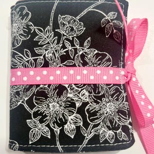 May include: A rectangular fabric cover featuring a black background with a white floral print. A bright pink ribbon with white polka dots is wrapped horizontally around the center and tied into a bow on the right side of the item.