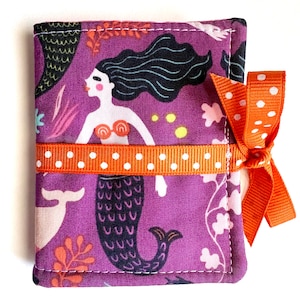 May include: A rectangular fabric case featuring a purple background with a repeating mermaid pattern, including black-haired mermaids, orange coral, and white sea life. The case is secured with an orange ribbon featuring white polka dots.