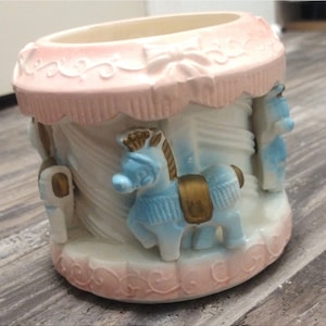 May include: A ceramic carousel-themed container in pink and white. It features light blue horses with gold accents. The top and base have a pink scalloped design, creating a whimsical aesthetic. A decorative item.